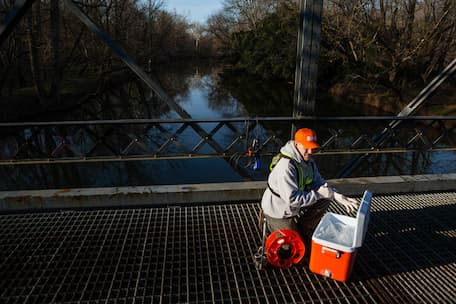 Male biologist kneels on a bridge above a creek preparing monitoring equipment.