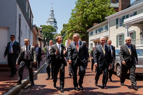 A group of male and female politicians walk down a brick street in downtown Annapolis with the city's capitol building in the background.
